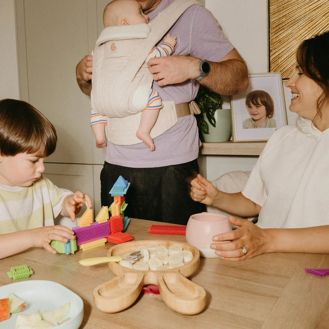Man holding a baby in a beige baby carrier looking down at a toddler playing at the table with a woman