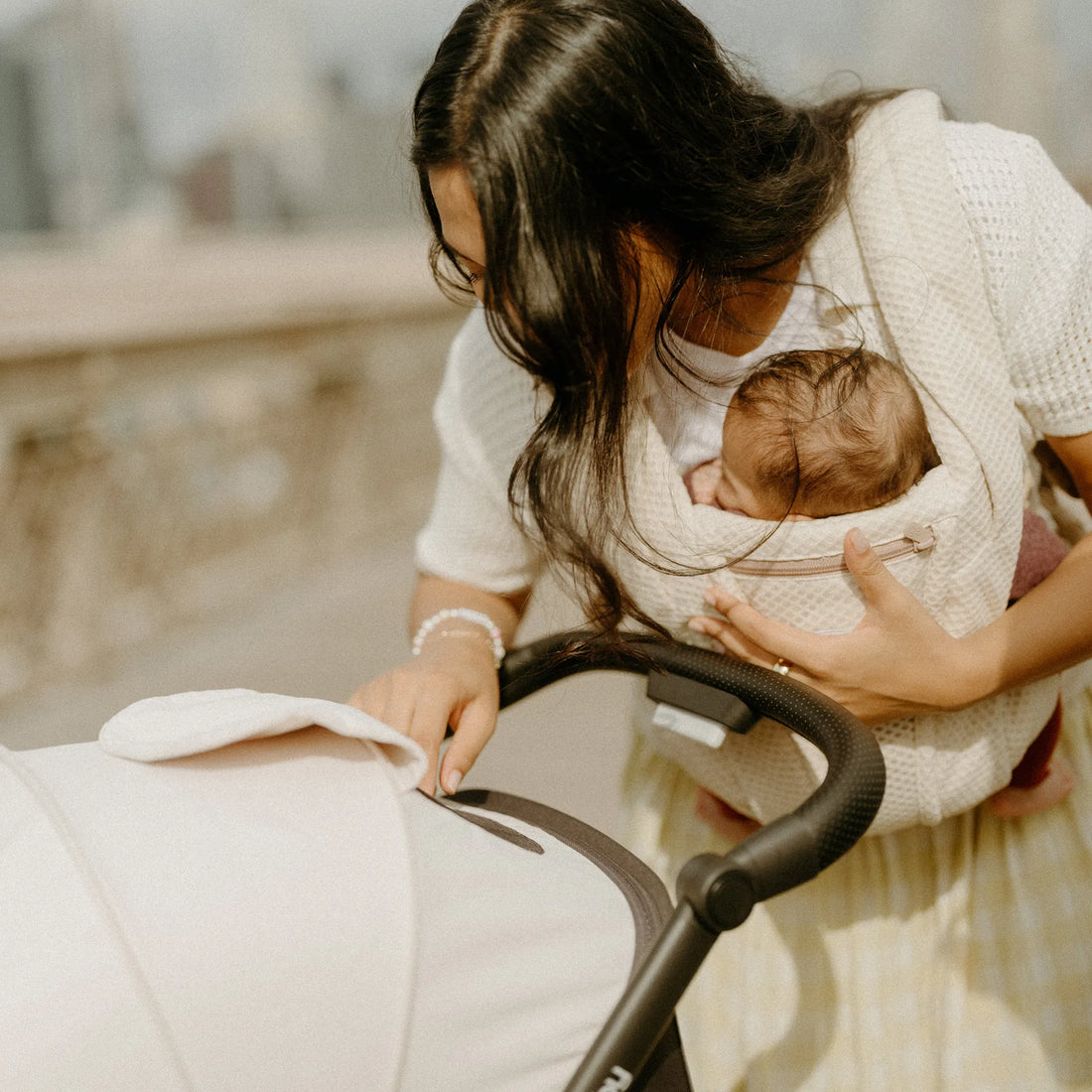 Woman holding a baby in a beige baby carrier looking down at a stroller