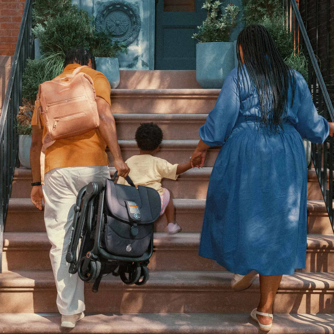 Woman holding hands with a toddler climbing the stairs with a man carrying a backpack and travel stroller