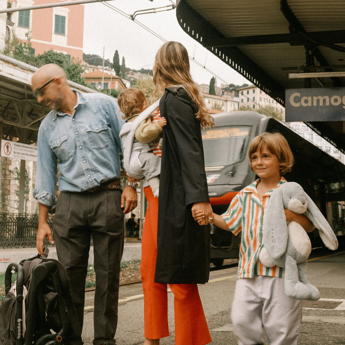 Woman holding a baby in a baby carrier holding hands with a toddler standing with a man holding a travel stroller