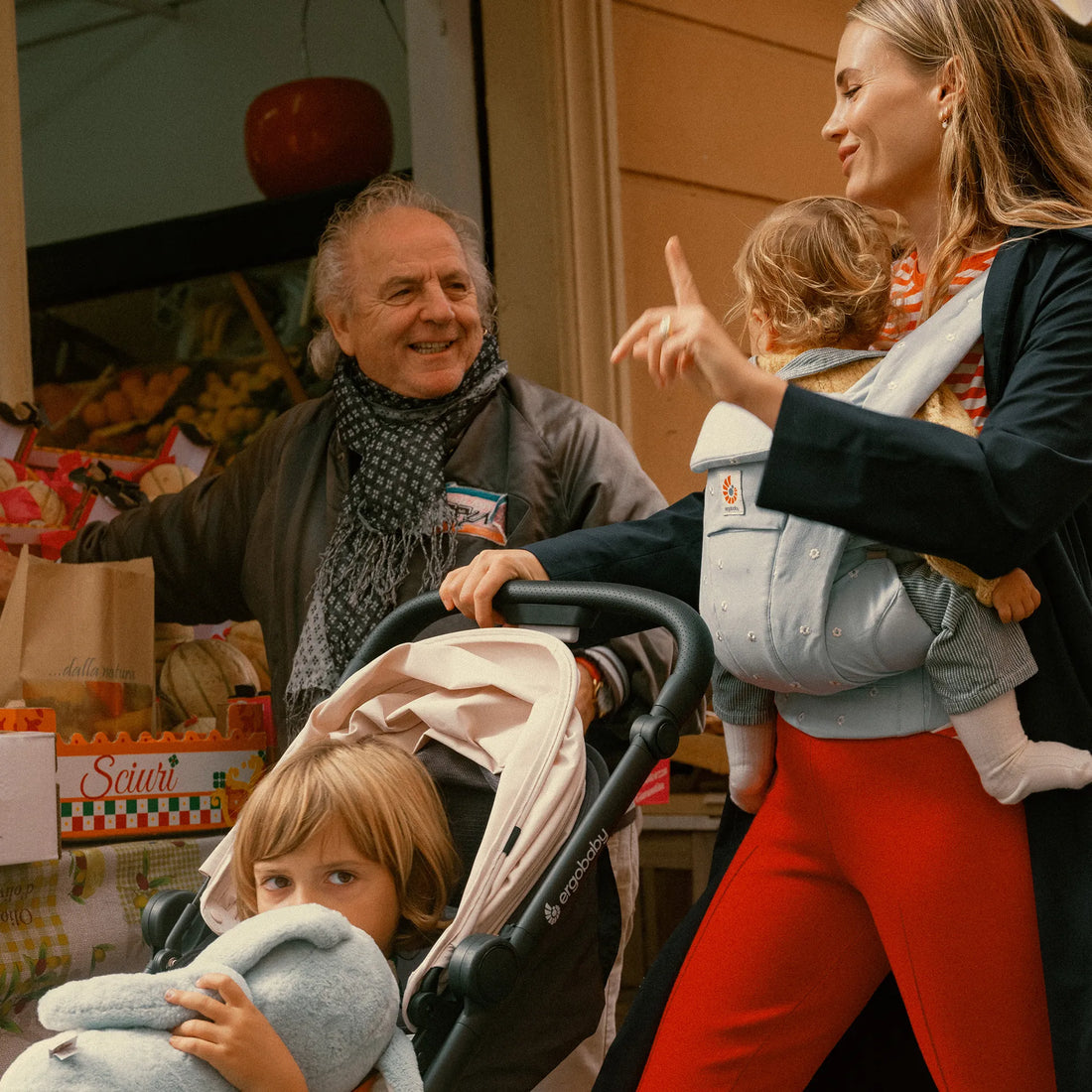 Woman holding a baby in a baby carrier pushing a toddler in a stroller while talking to a man