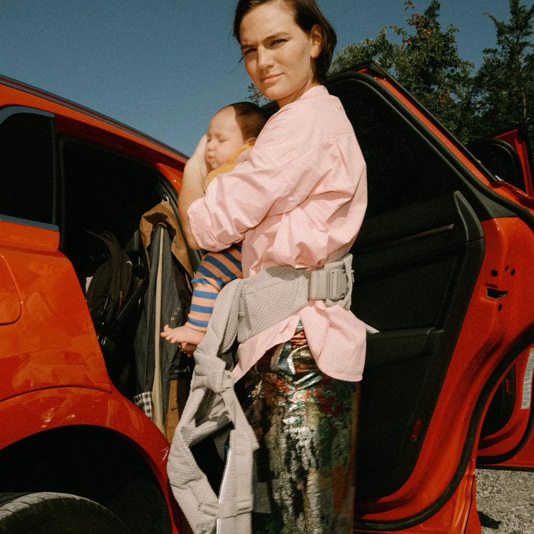 Woman holding a baby in a grey baby carrier standing outside a car
