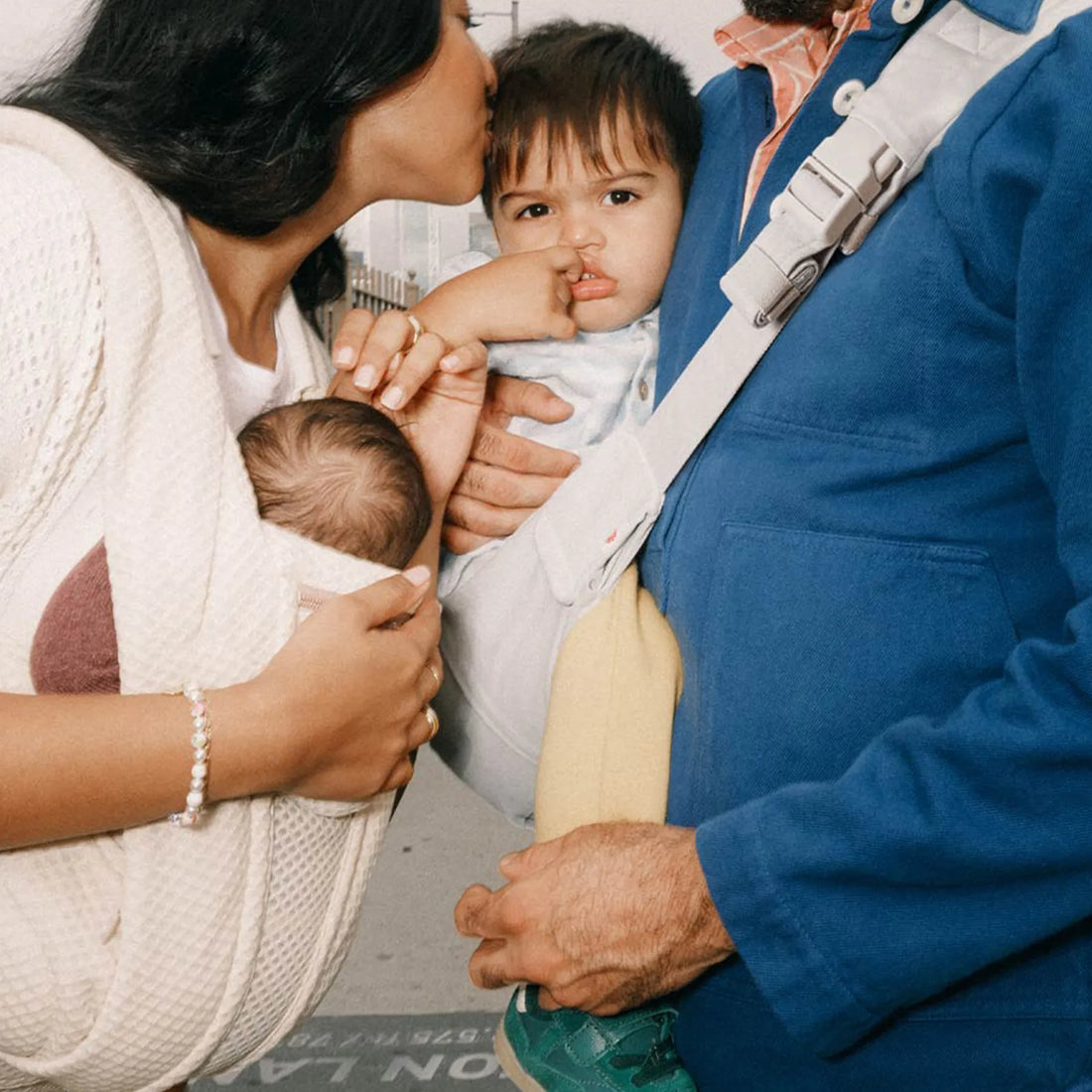 Woman holding a baby in a beige baby carrier alongside a man holding a toddler in a grey sling