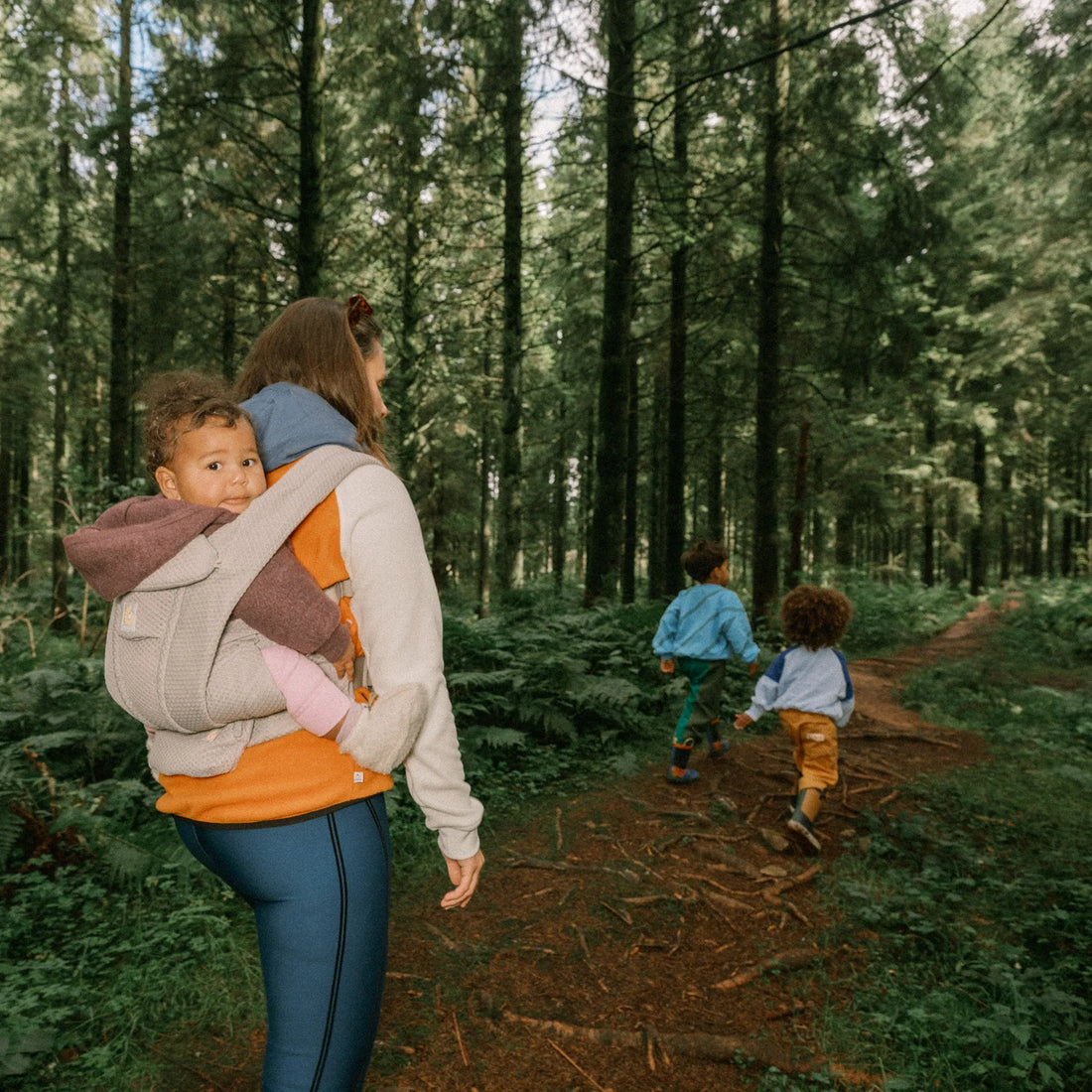 Woman holding a baby on her back in a beige baby carrier following two toddlers