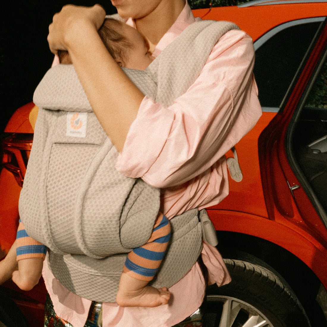 Woman holding a baby in a grey baby carrier in front of a car