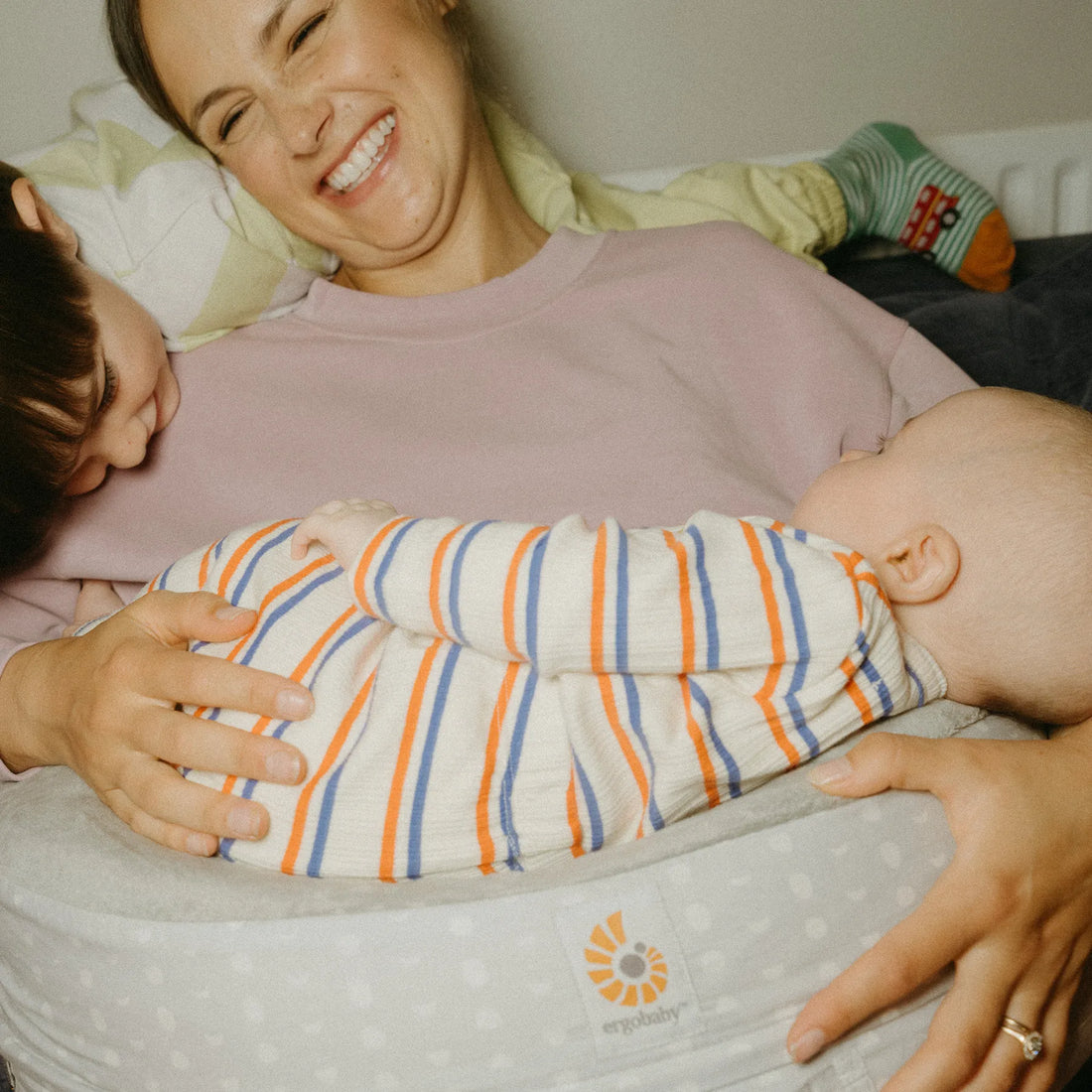 Woman lying down with a baby wrapped in her arms as toddler looks on