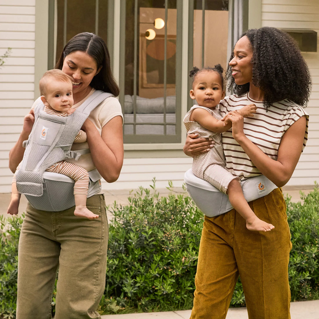 One women holding a baby in a grey baby carrier walking with a women holding a baby on her hip in a hip seat