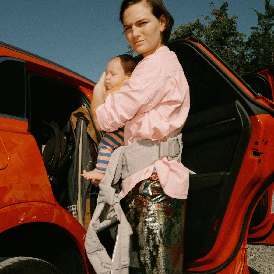 Woman holding a baby in a baby carrier next to a car