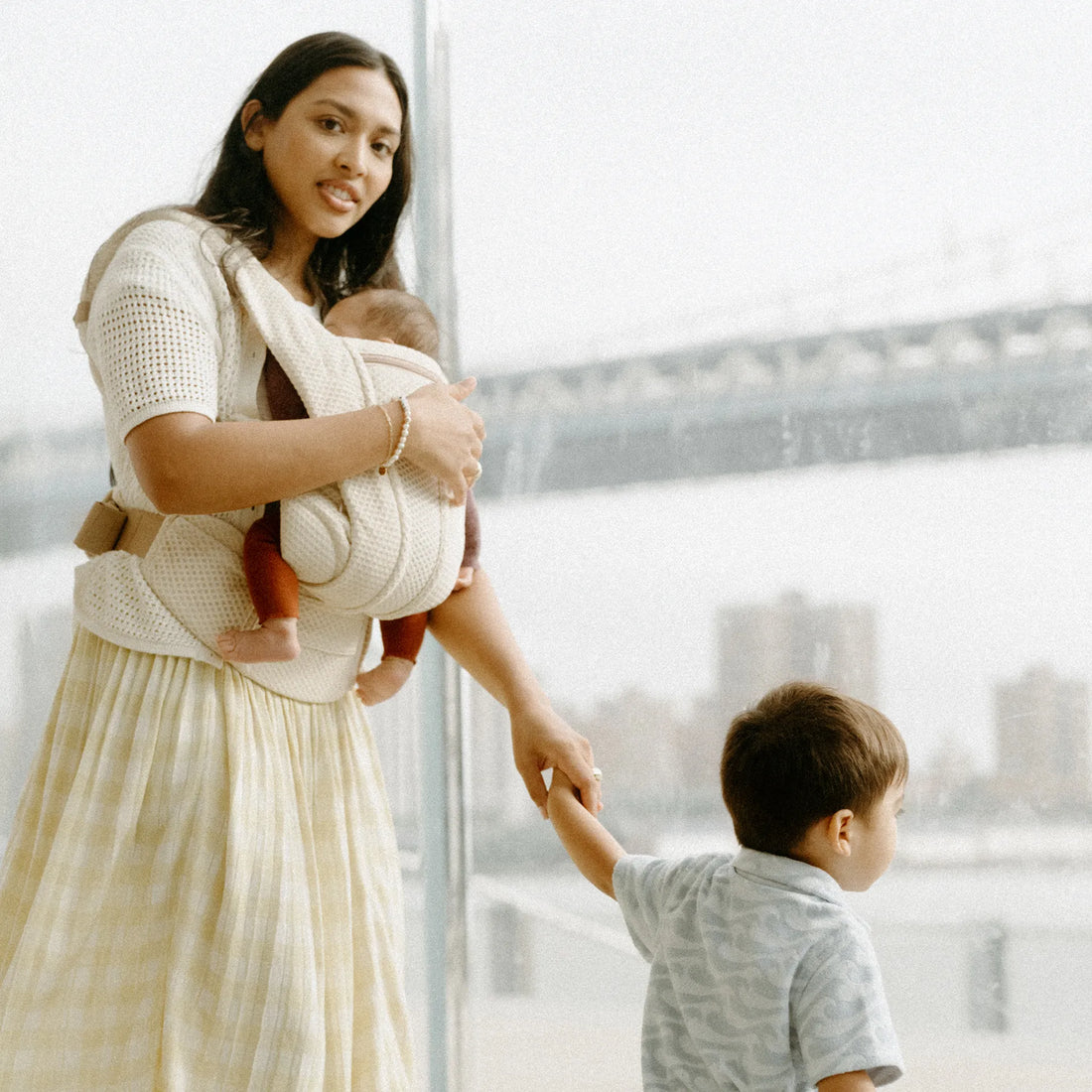 Woman holding a baby in a white baby carrier and holding a toddlers hand