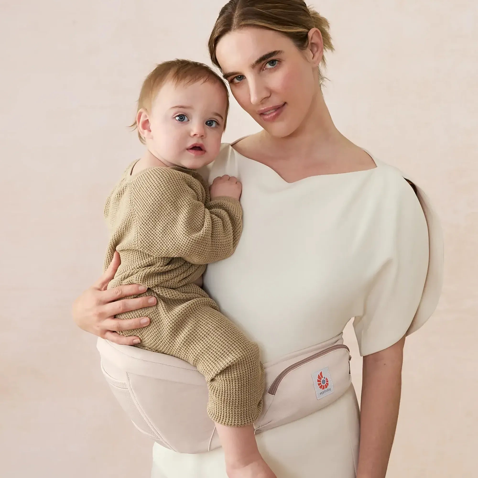 Woman holding a baby in a beige ergobaby baby carrier against a light background | Ergobaby United States