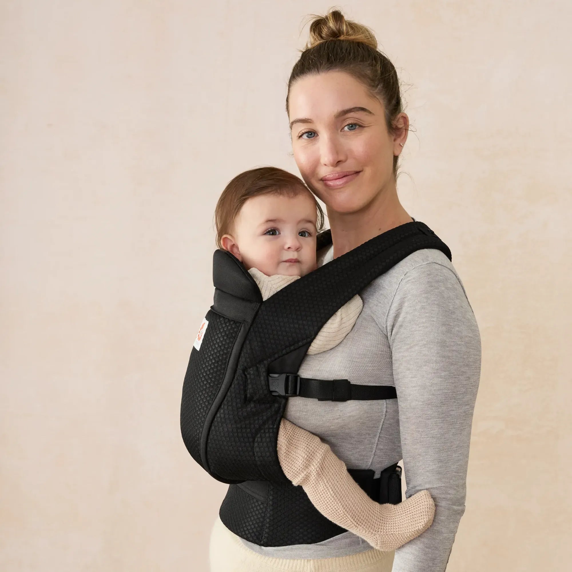 Woman holding a baby in a black mesh omni lite baby carrier against a beige background | Ergobaby Spain
