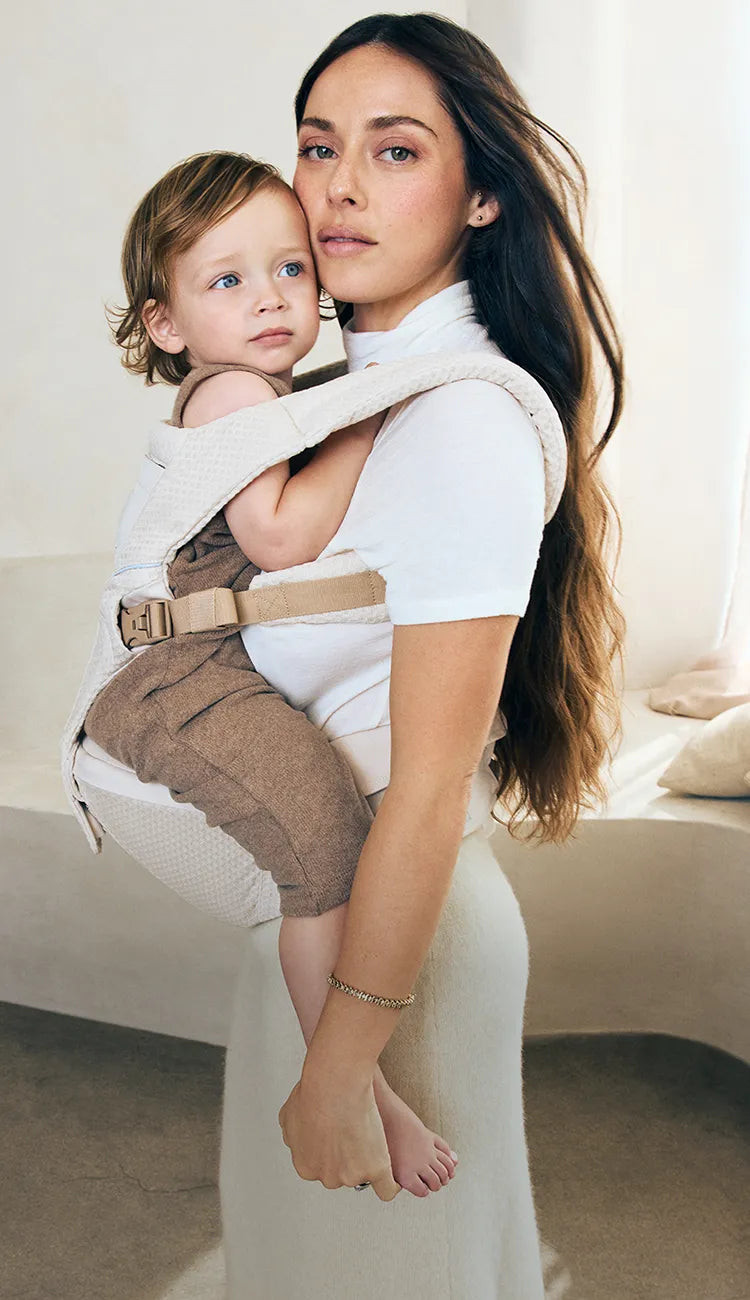 Woman holding a child in an ergobaby alta baby carrier against a white background