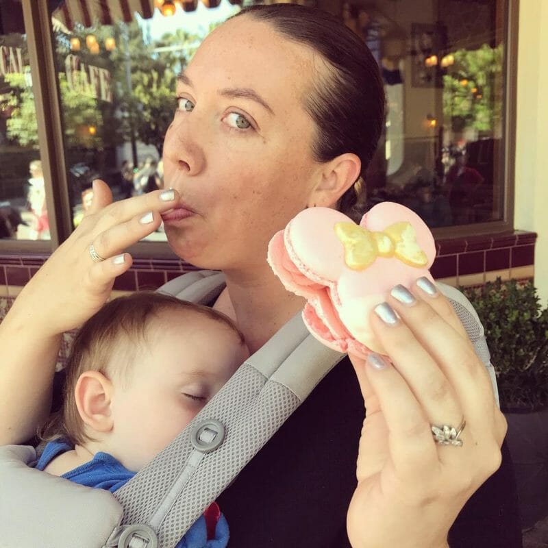 A mom enjoys a Mickey cookie while baby sleeps in an Ergobaby baby carrier at Disneyland